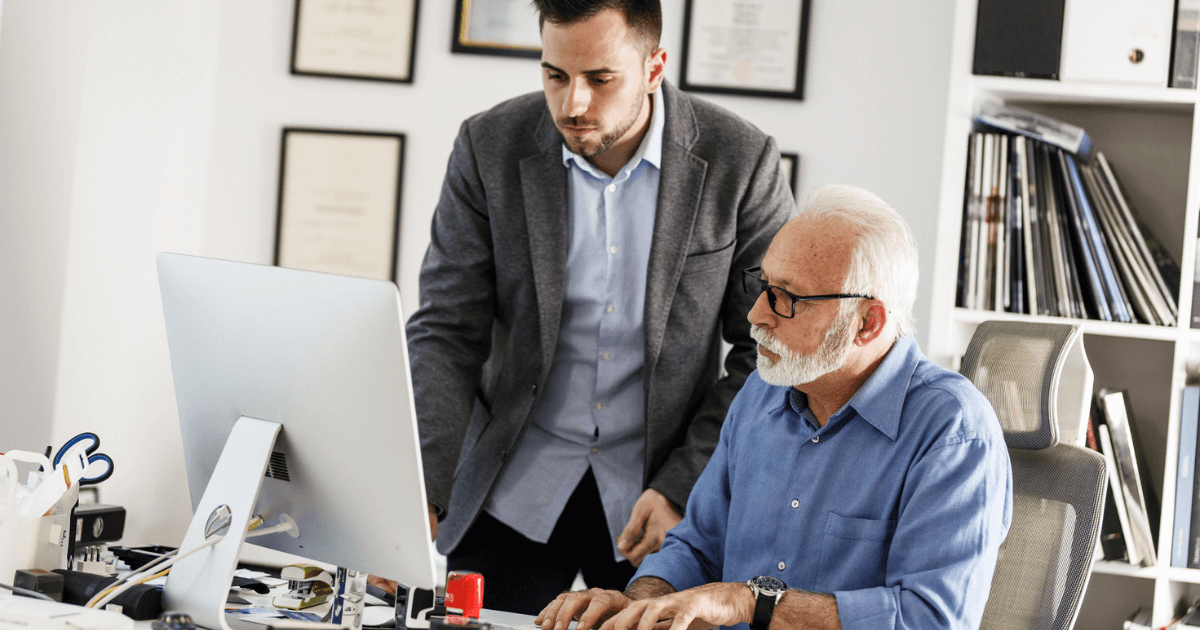 Adult man standing over senior man at desk with computer reviewing reentering the workforce.